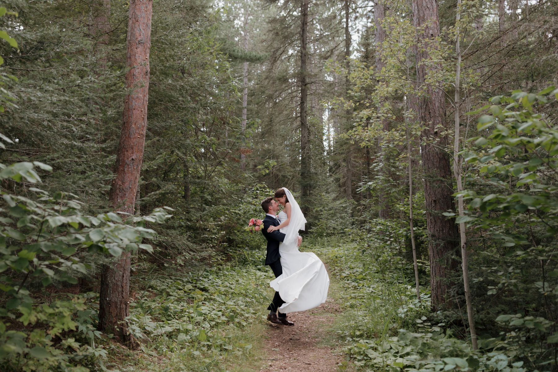 a couple taking wedding photos on a trail the groom is lifting the bride up in the air 