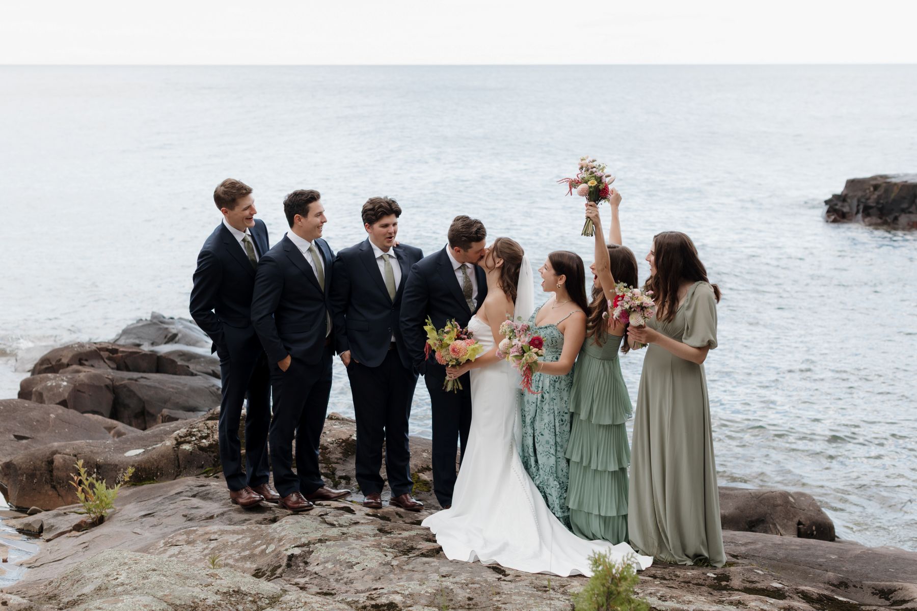 a couple with their wedding party taking photos next to the water the bride and groom and kissing 