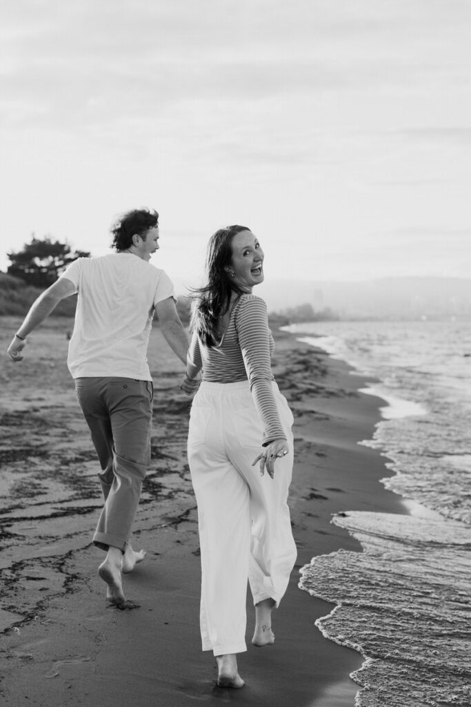 A couple holding hands and running on the beach as one looks over their shoulder 
