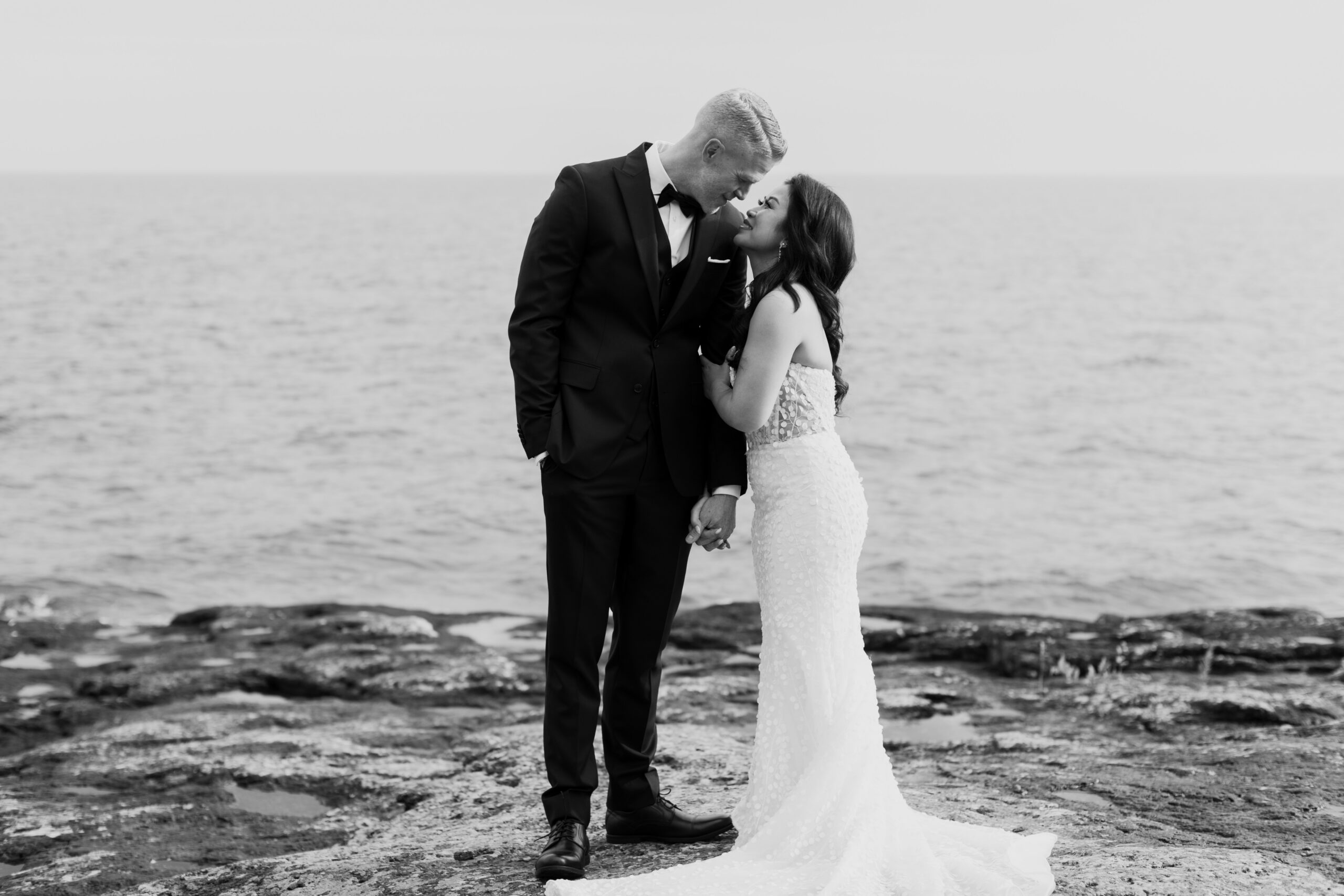 a couple taking wedding portraits next to Lake Superior the woman is holding her partner's arm and they are leaning towards each other 