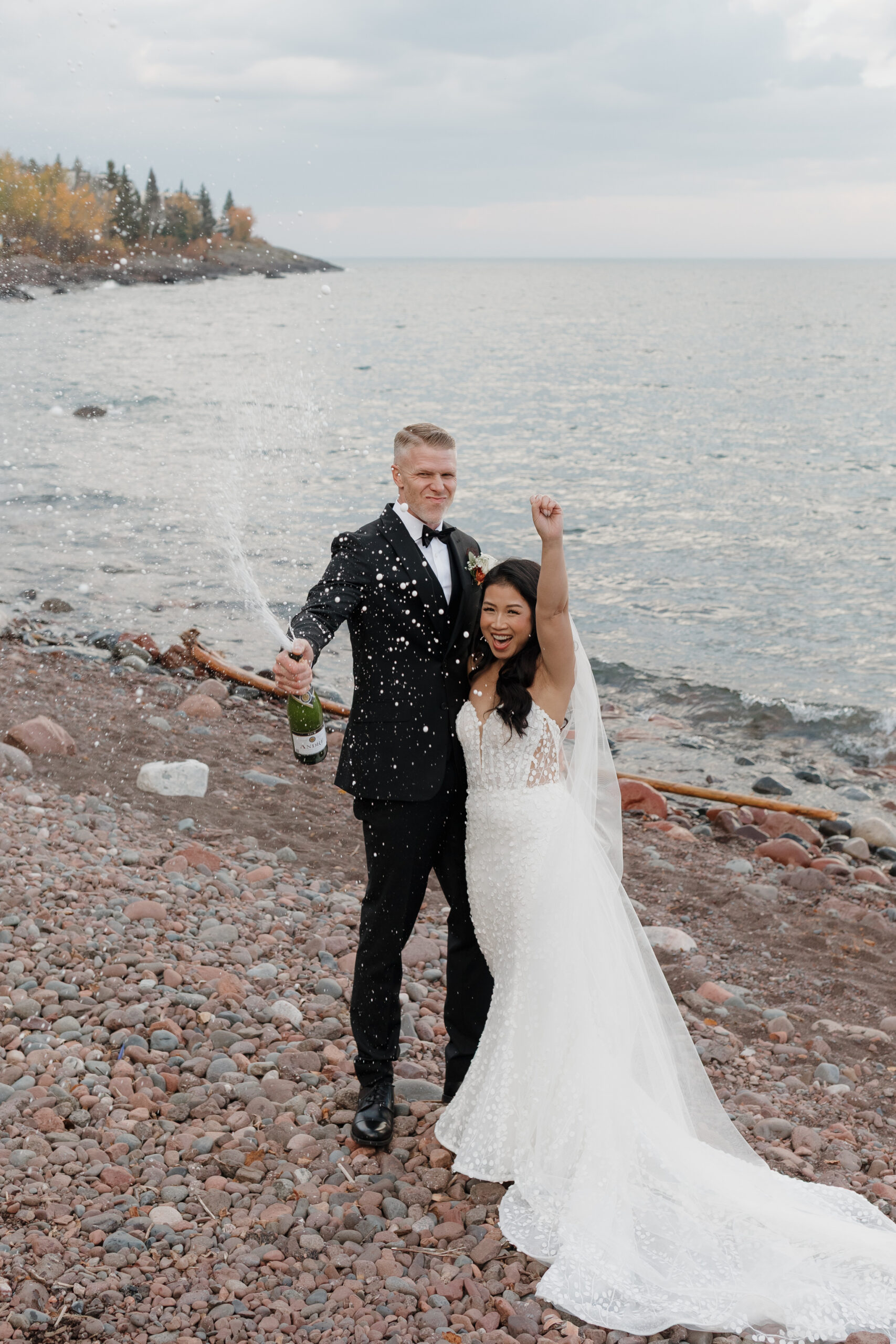 a couple popping a bottle of champagne on the day of their wedding next to lake superior 
