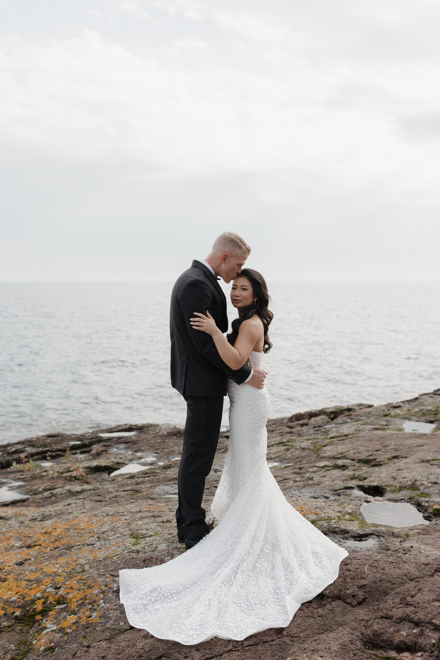 a couple taking wedding portraits next to lake superior the groom is hugging his partner and kissing her forehead 